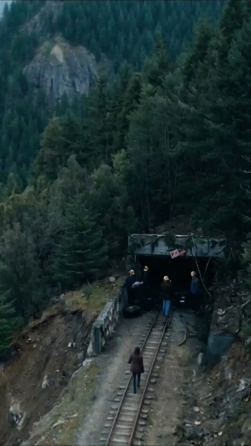 A still from Dark Hollow: people walk along rail tracks to a mine entrance in a forest